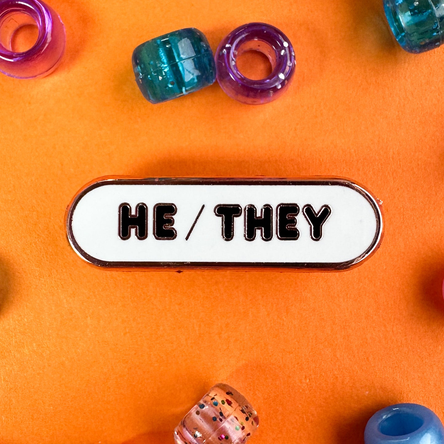 A white capsule shaped pin to share your pronouns, it has "He/They" in bubble letters. The pin is on an orange background with pony beads around it.