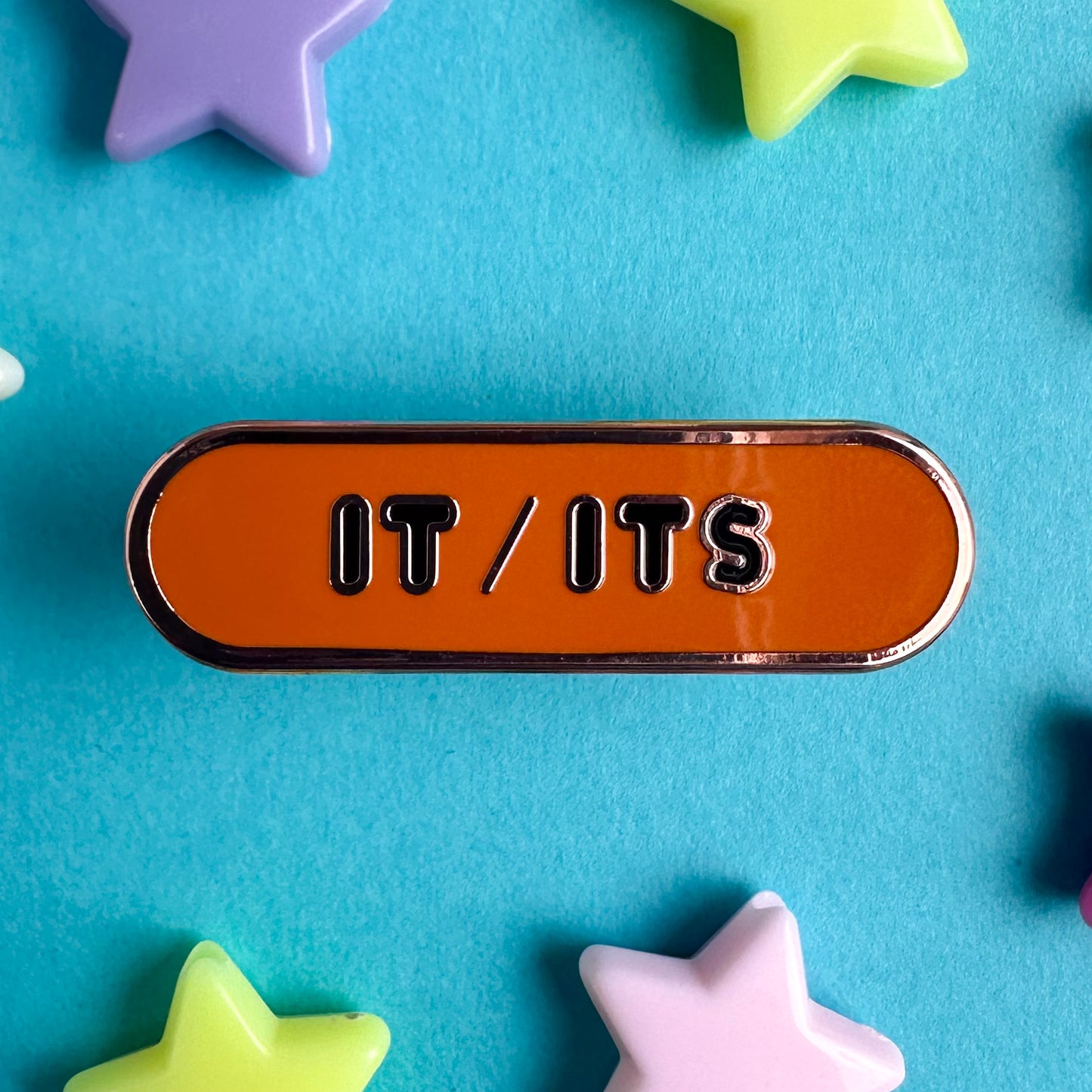 An orange oval shaped enamel pin with the words "It/Its" on it in black bubble letters. The pin is sitting on a blue piece of paper with plastic star beads around it.