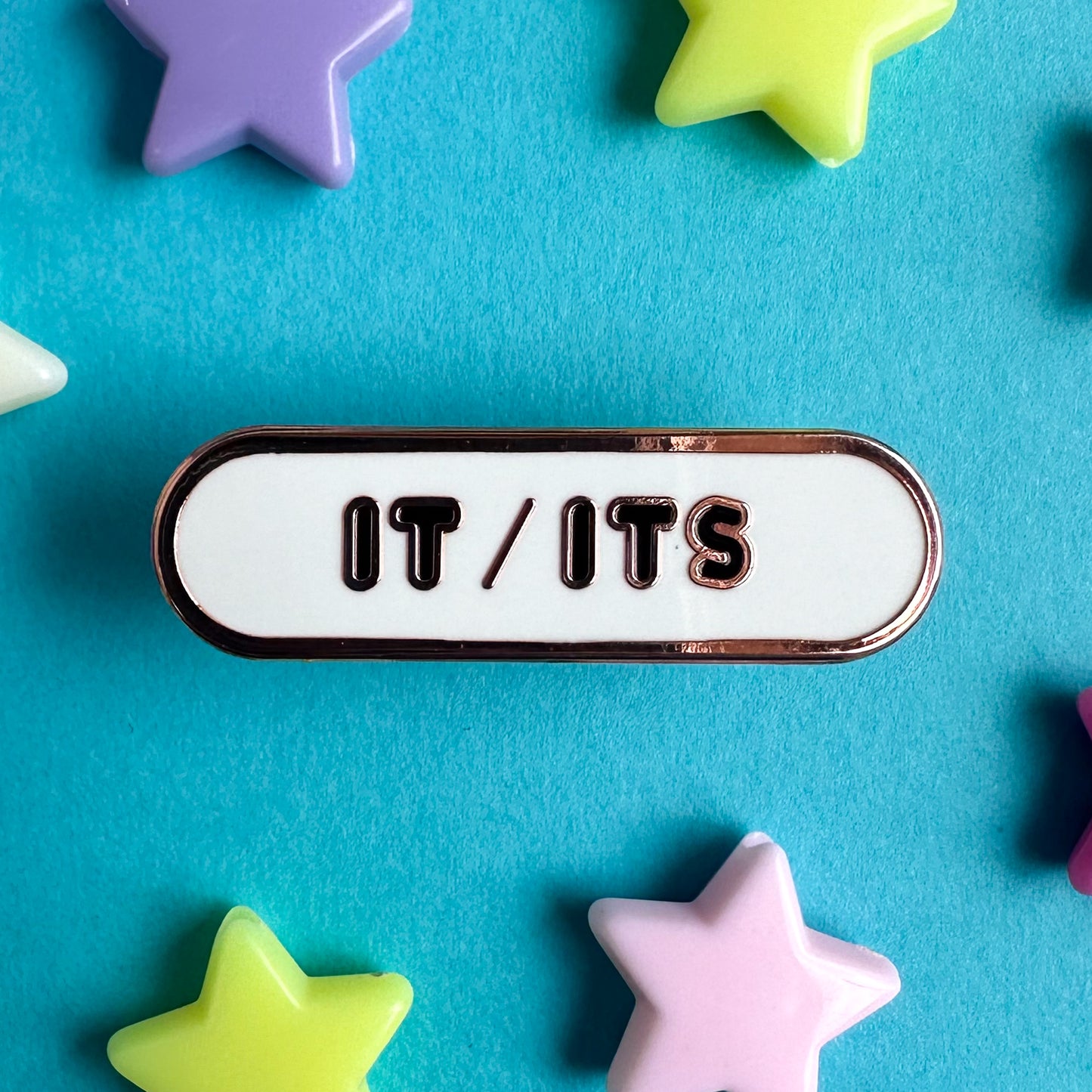 A white oval shaped pin with black bubble letters that read "it/its". The pin is sitting on a blue piece of paper with plastic star beads around it.