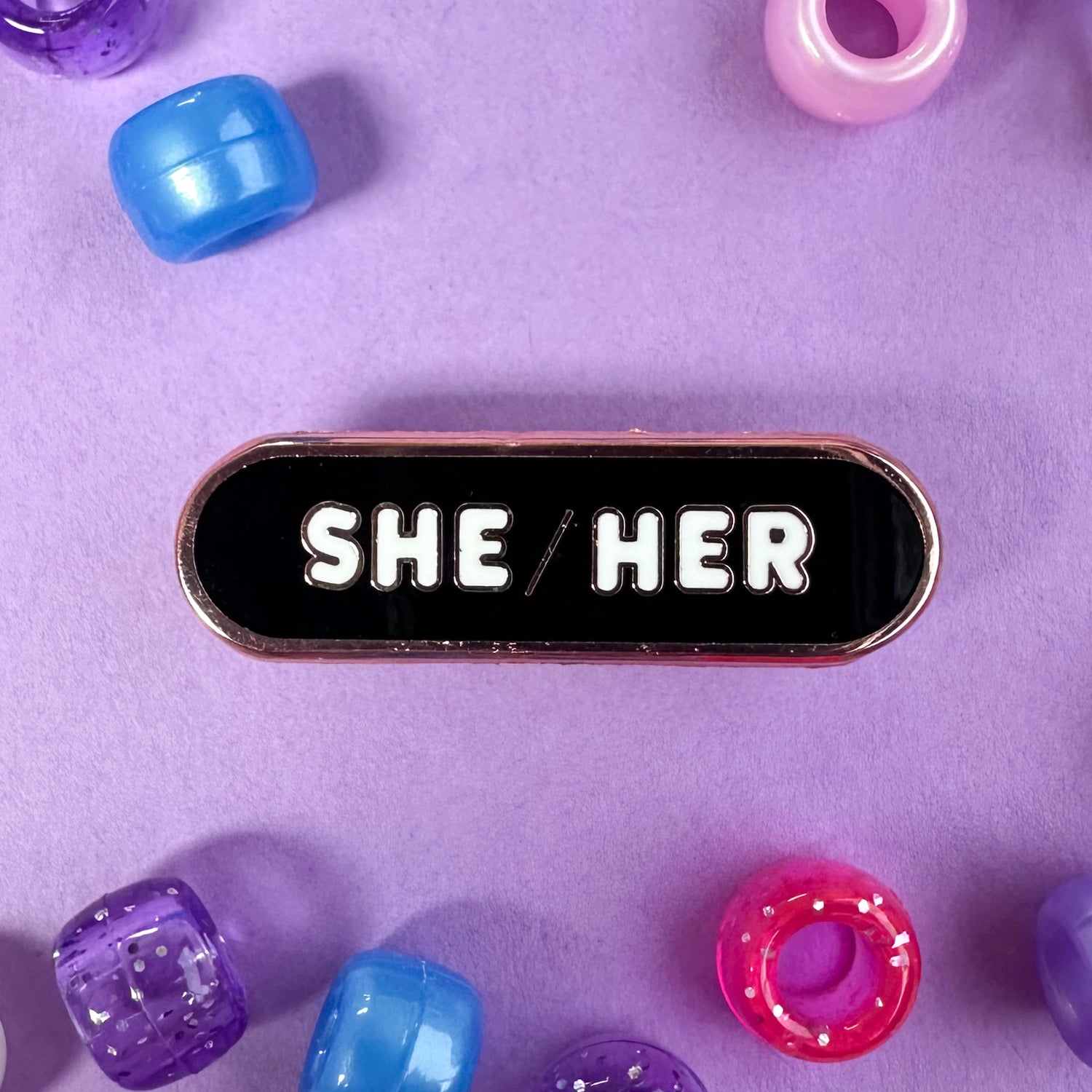 A capsule shaped pin with the pronouns "She/Her" on it. The pin is on a lavender background with blue, purple, and pink pony beads scattered around it.