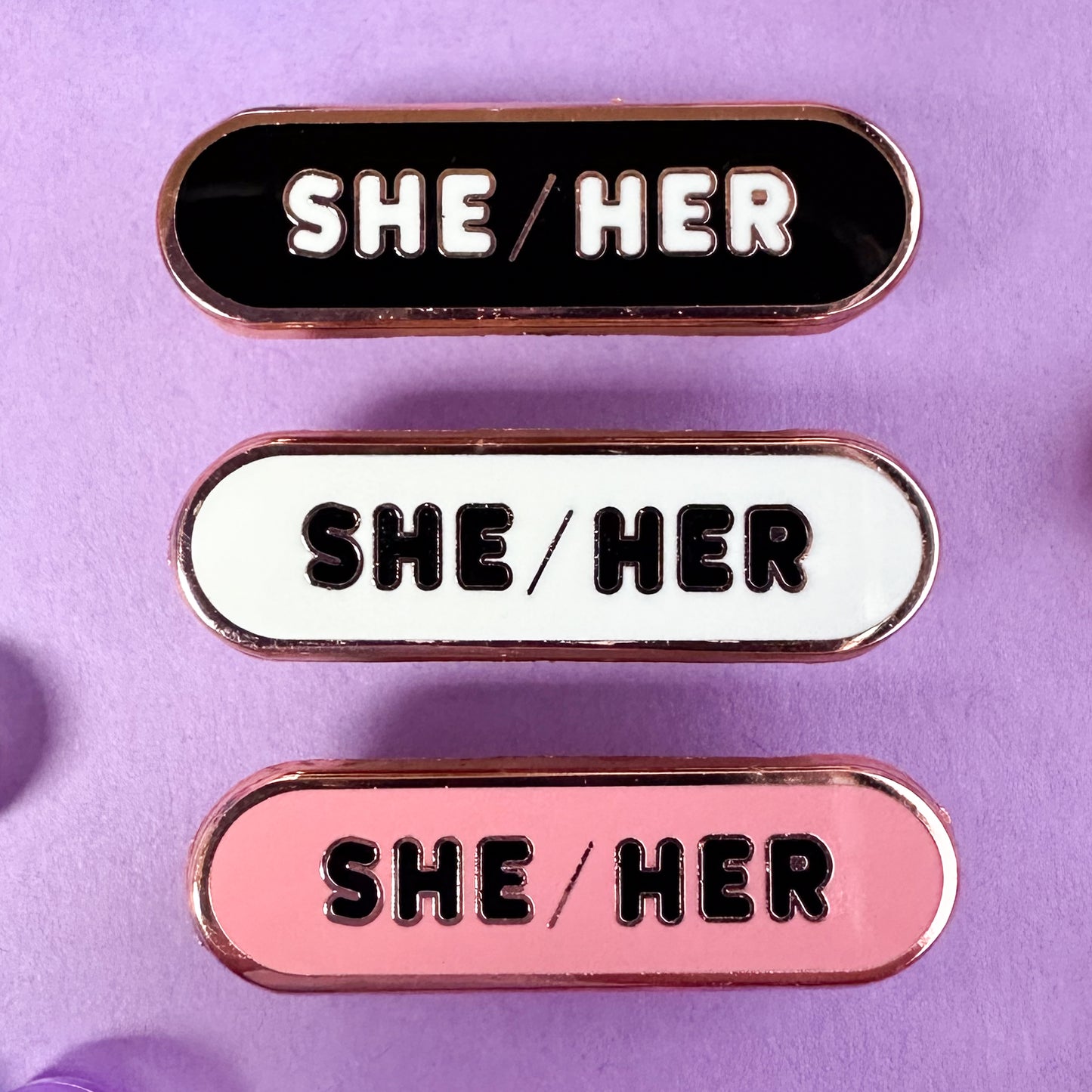 Three oval shaped pins with the words "She/Her" on them, one pin is black, one is white, and one is pink. The pins are on a lavender background.