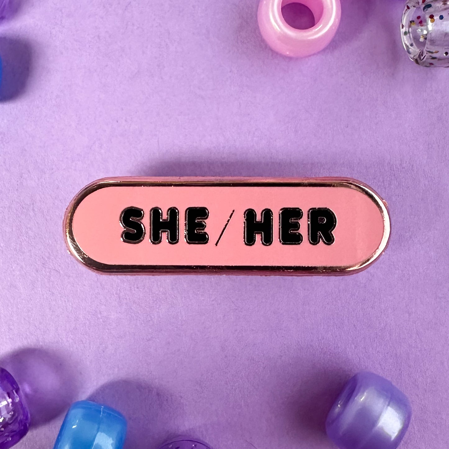 A pink enamel pin with the words "She/Her" on it. The pin is on a lavender background with pink, blue, and purple pony bead around it.