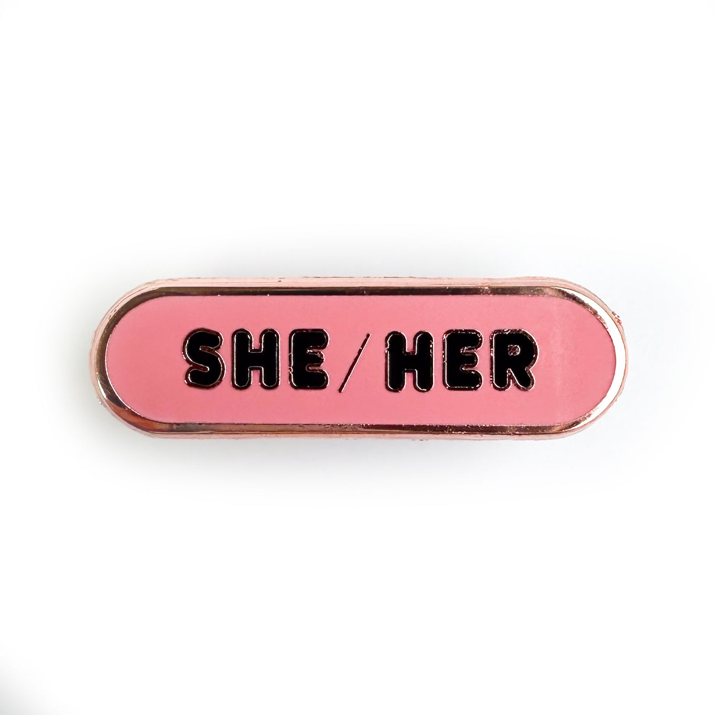 A pink enamel pin with the words "She/Her" written on it in black letters.