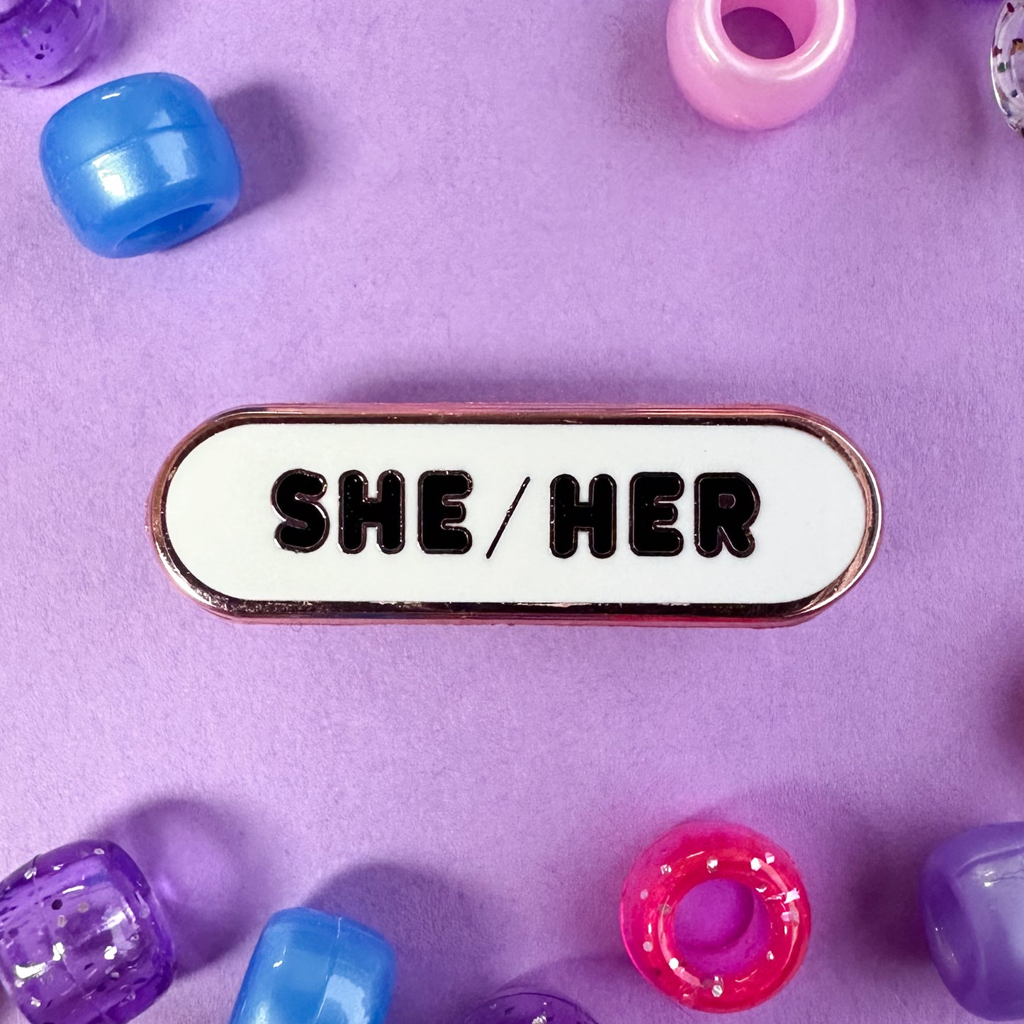A capsule shaped pin with the pronouns "She/Her" on it. The pin is on a lavender background with pink, purple, and blue pony beads scattered around.