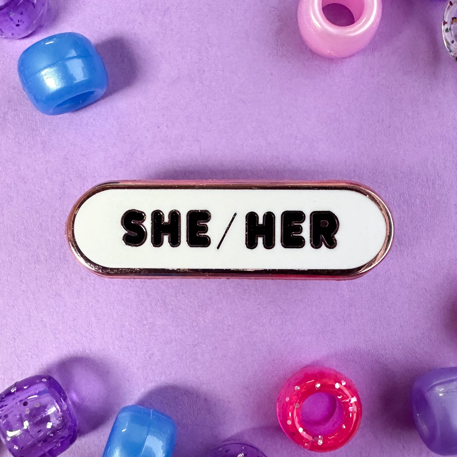 A capsule shaped pin with the pronouns "She/Her" on it. The pin is on a lavender background with pink, purple, and blue pony beads scattered around.