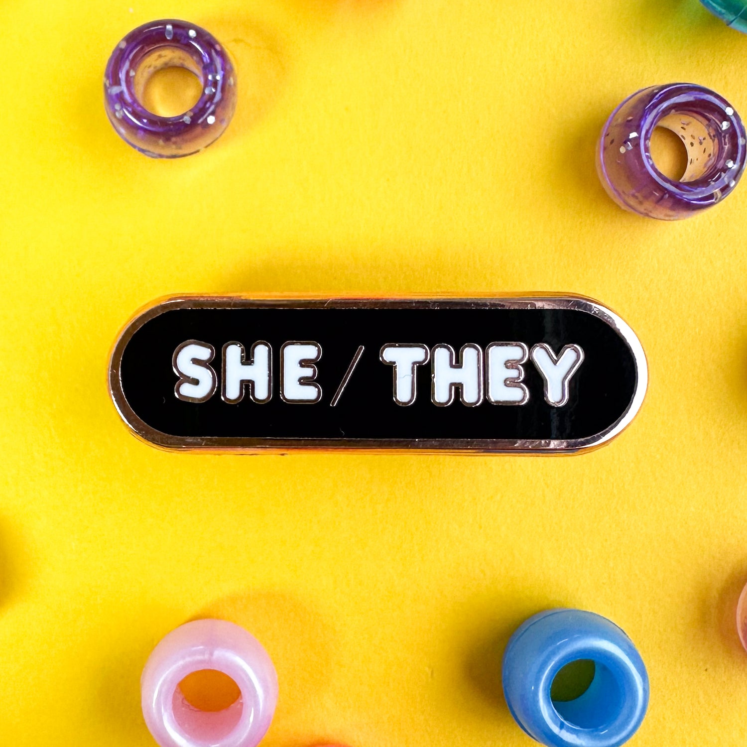 An oval shaped pin with the pronouns "She/They" on it. The pin is on a yellow background with purple, blue and pink pony beads around it.