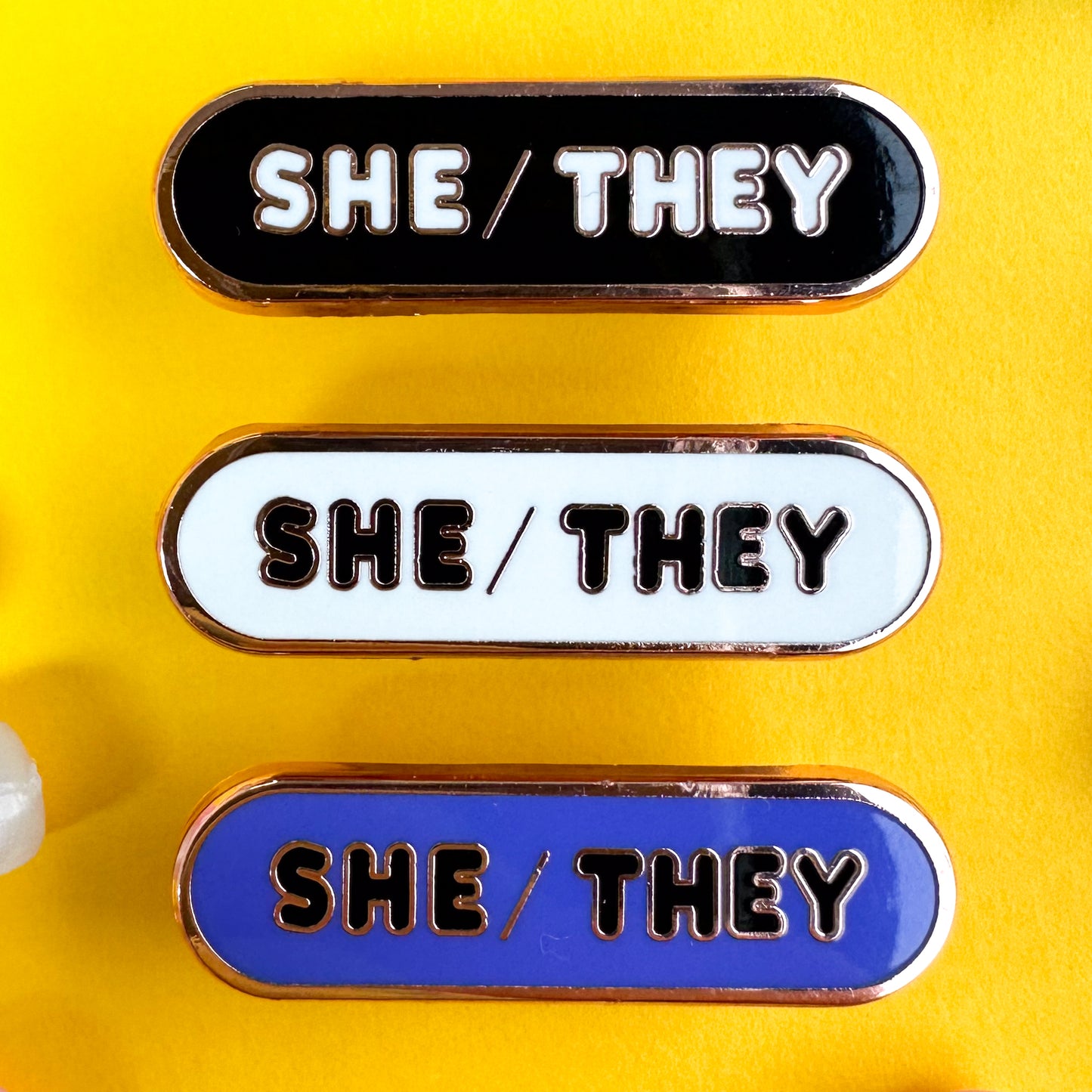 Three capsule shaped pins that have the words "She/They" on them one is black, one is white, and one is indigo. The pins are on a yellow background.