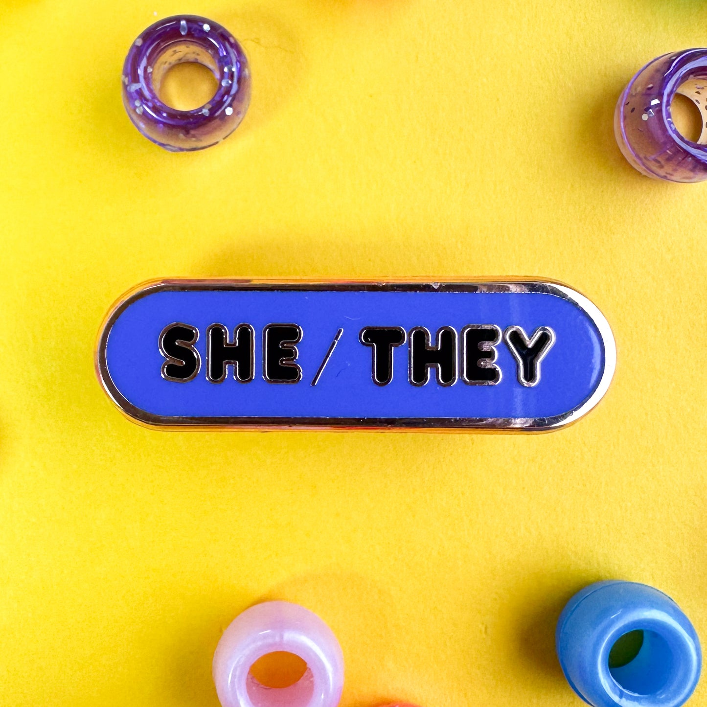 A capsule pin with black letters that read "She/They" on an indigo background. The pin is on a yellow background with blue, pink, and purple pony beads around it.
