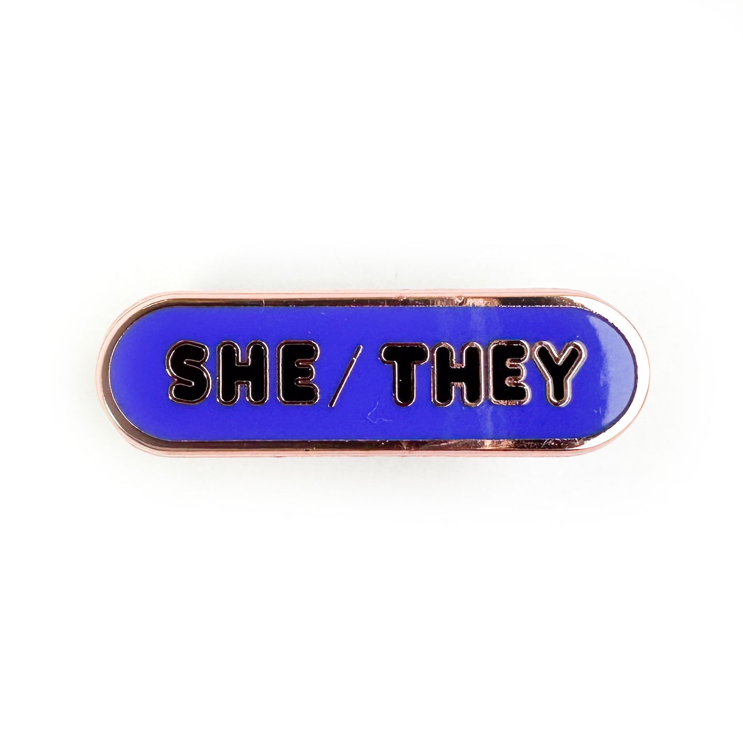 An indigo oval shaped pin with bubble letters on it that spell "She/They" on it.