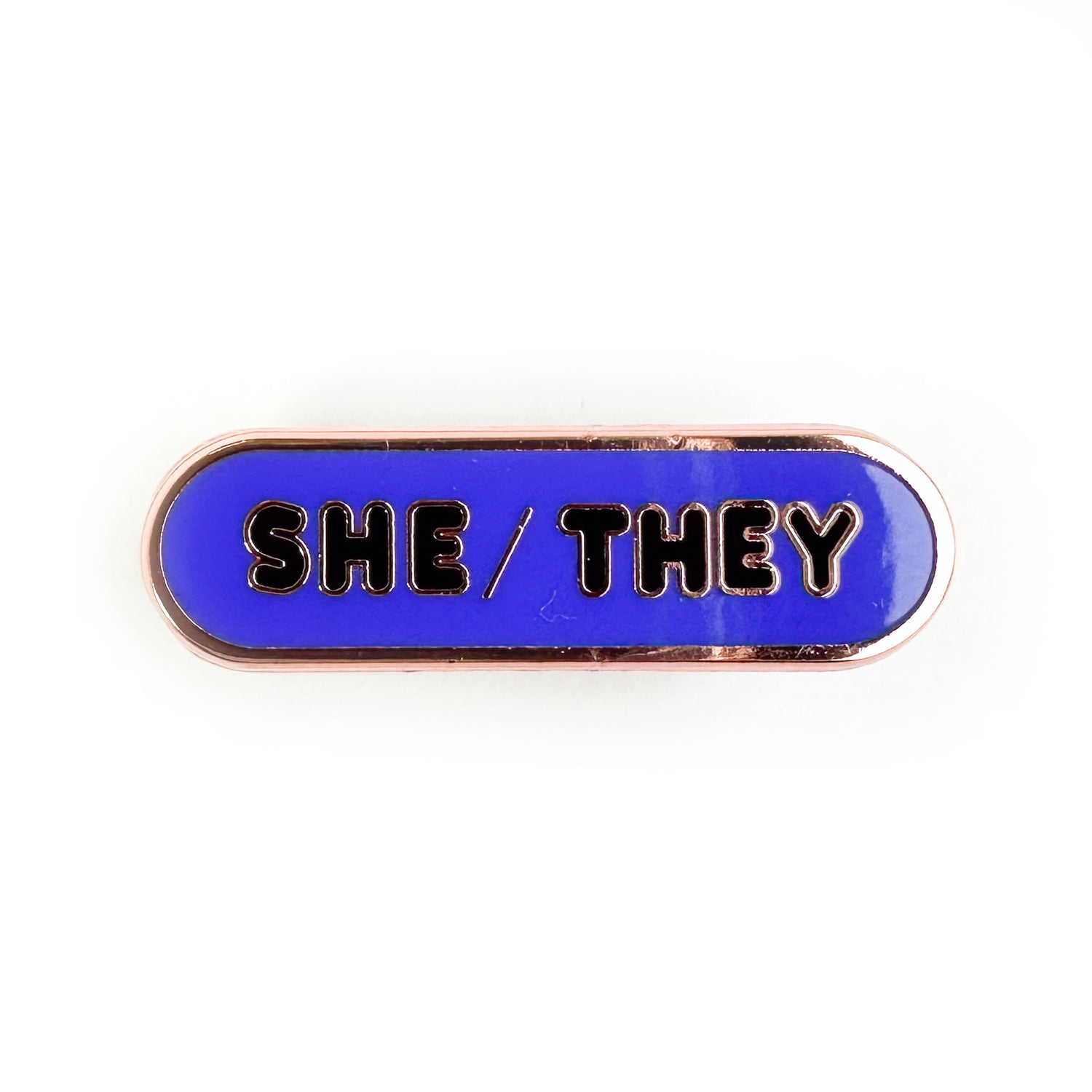 An indigo oval shaped pin with bubble letters on it that spell "She/They" on it.