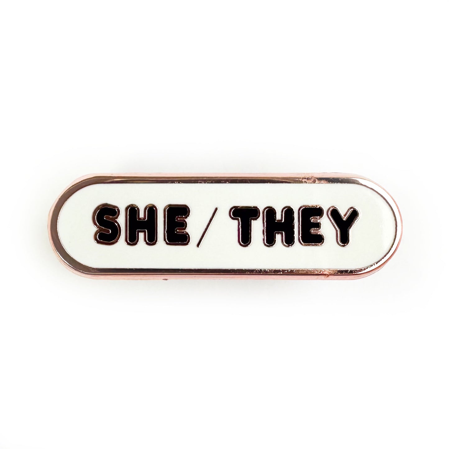 A pin with bubble letters that spell the pronouns "She/They" on it.