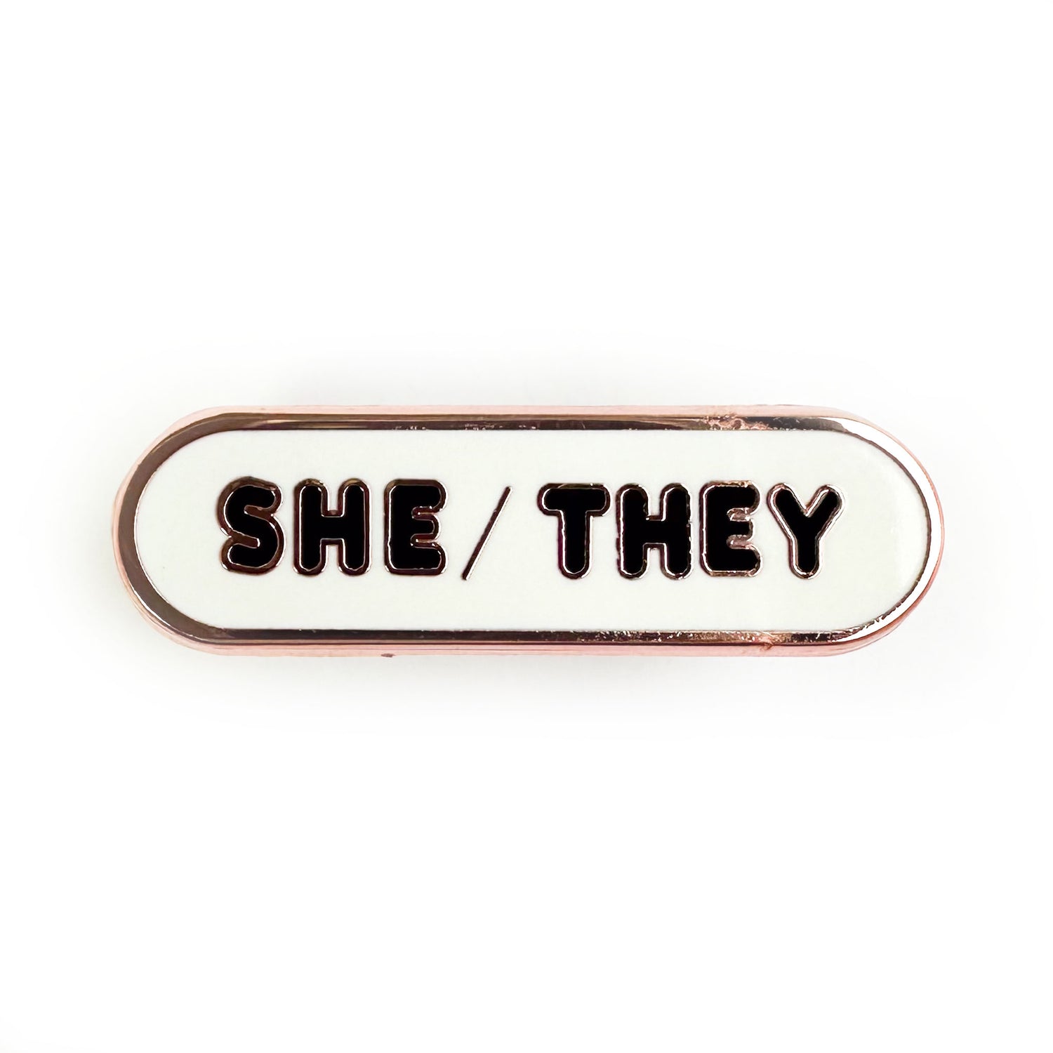 A pin with bubble letters that spell the pronouns "She/They" on it.