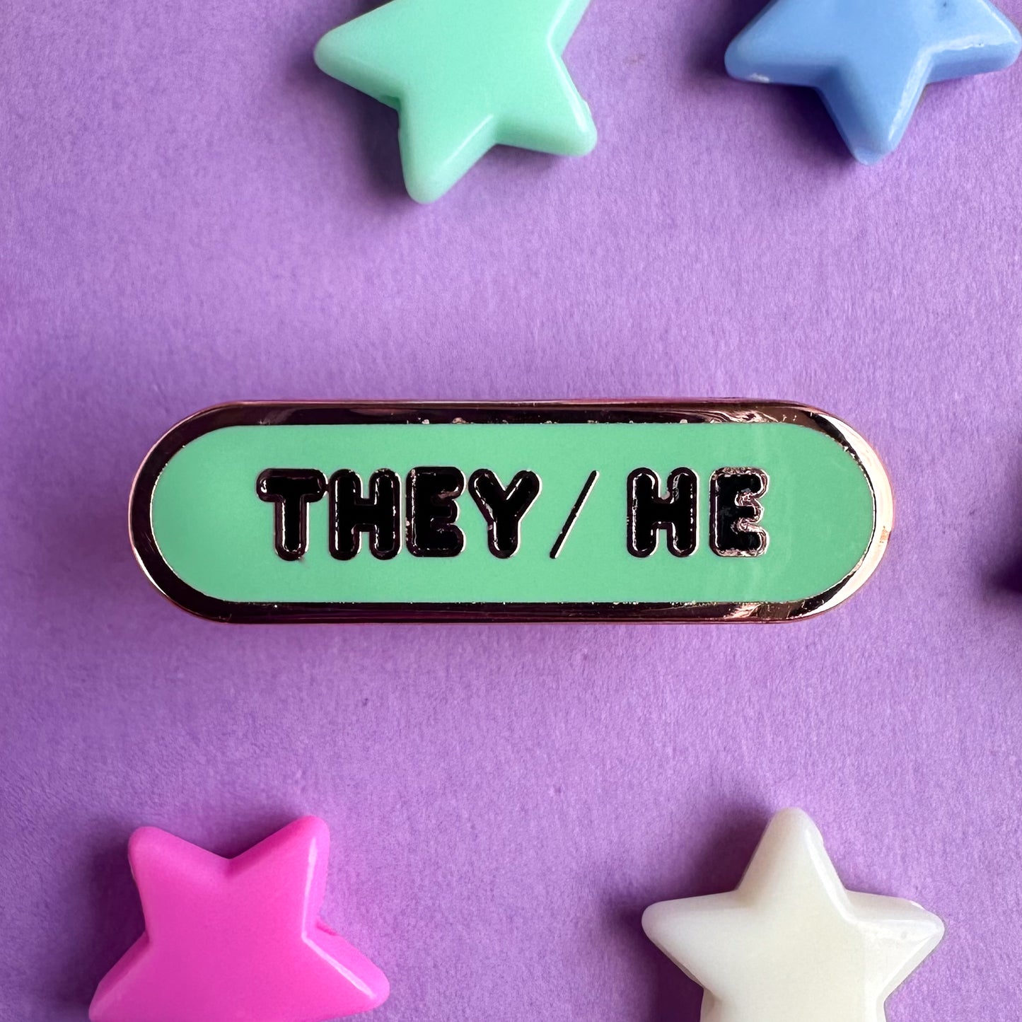 A green pin shaped like a bandaid with the pronouns They/He on it in black bubble letters. The pin is on a purple background with plastic star beads around it.