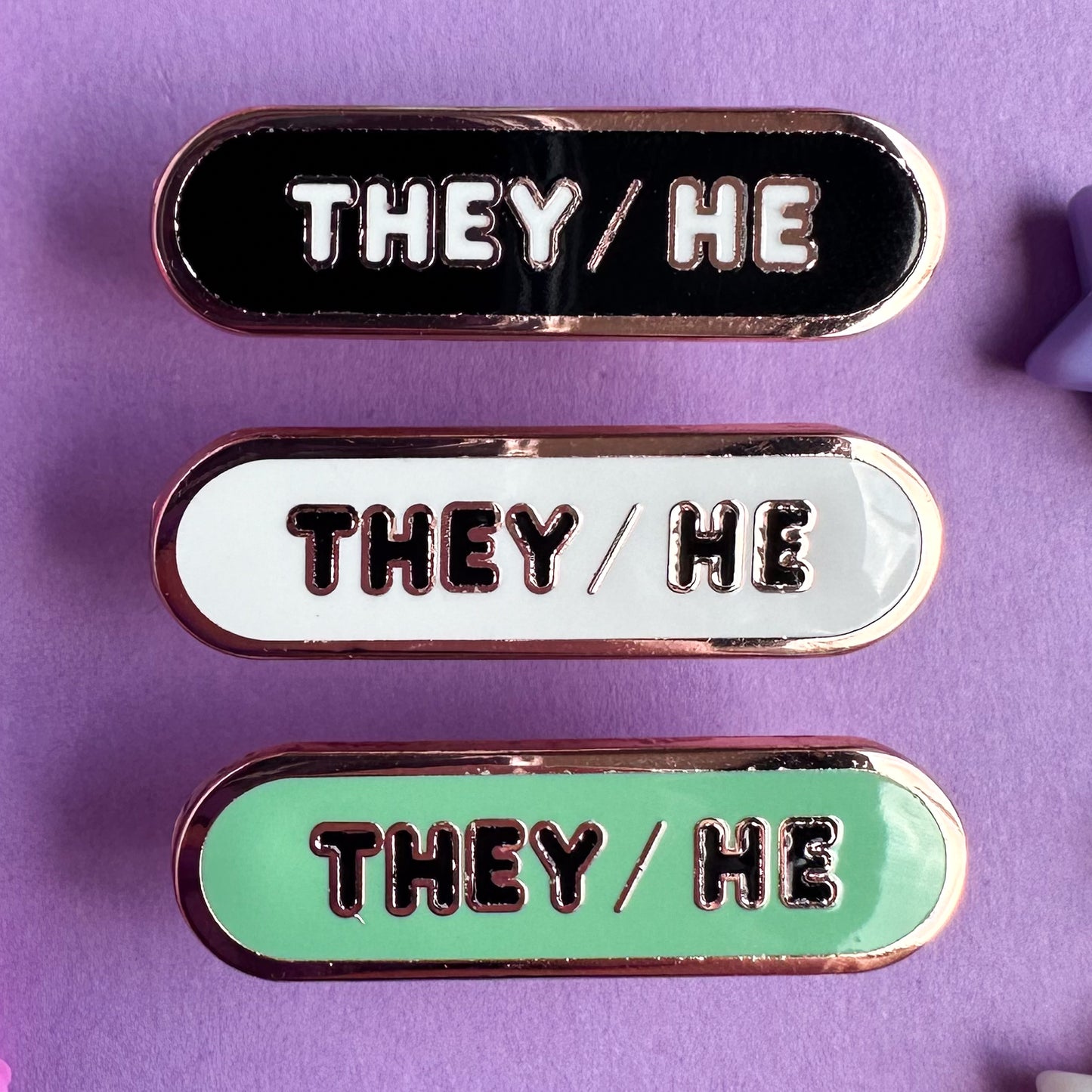 Three capsule shaped enamel pins with the words "They/He" on them, the top pin is black, the middle is white, and the bottom one is green. The pins are on a purple background.