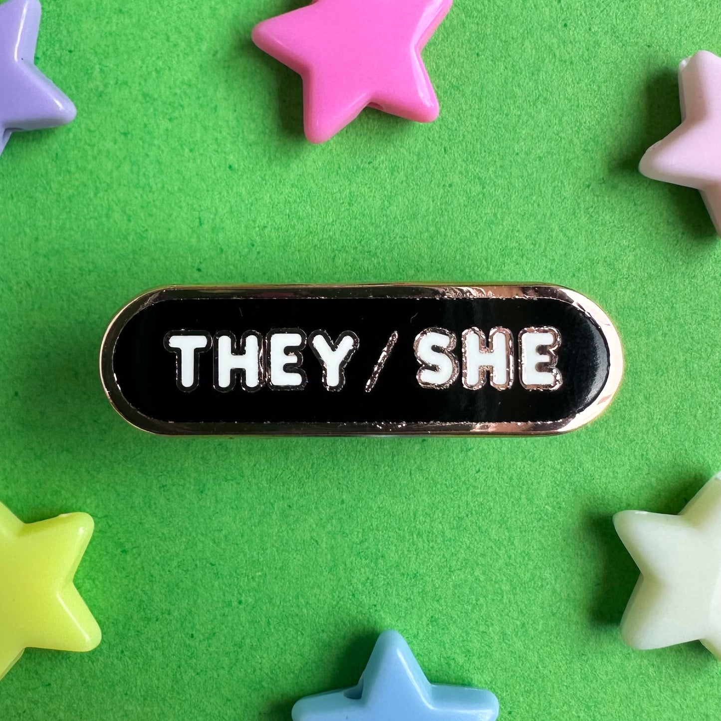 A capsule shaped enamel pin with white bubble letters that spell "They/She" the pin is on a green background with pastel stars around it.