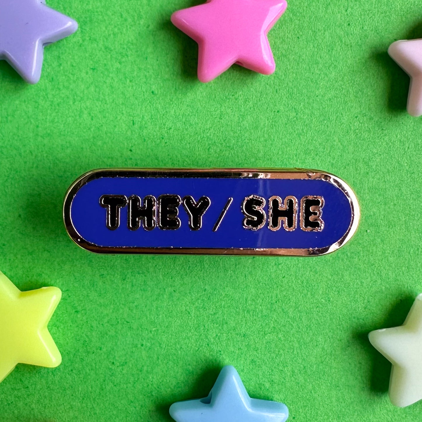 An indigo oval shaped enamel pin with the words "They/She" in black bubble letters. The pin is on a green background with plastic star beads around it.