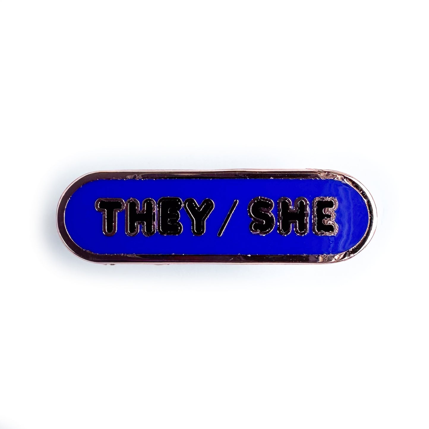 A capsule shaped enamel pin that is indigo with black letters "They/She" on it.