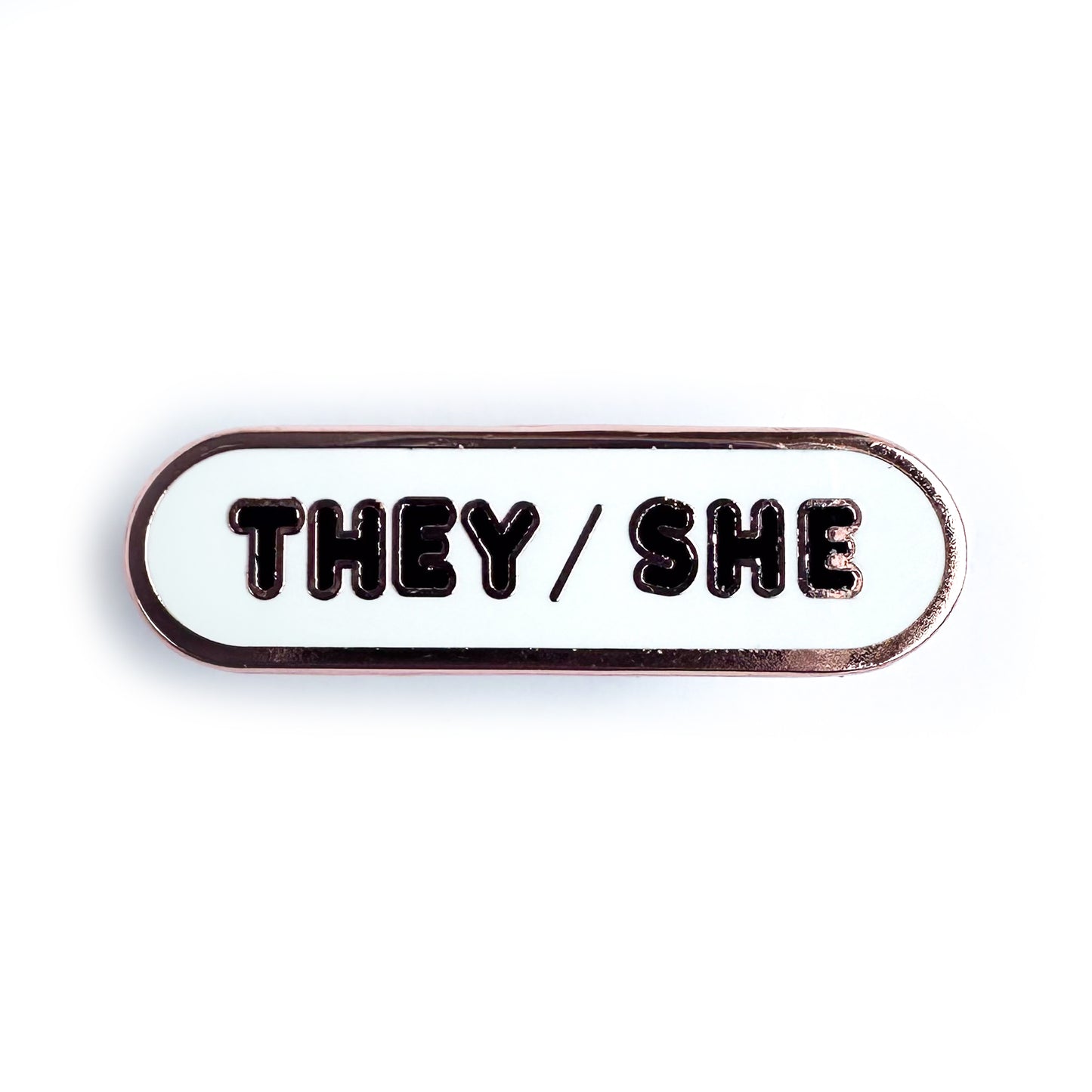 A white bandaid shaped enamel pin with black bubble letters that spell "They/She" on it.
