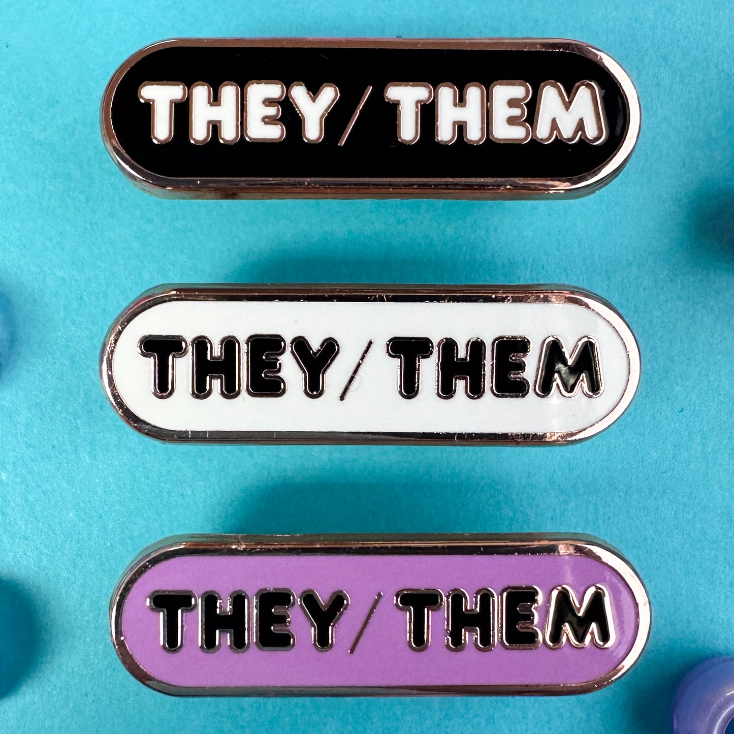 Three pins with the pronouns "They/Them" written on them, one pins is black, one is white, and one is lavender. The pins are on a blue background with blue pony beads around it.