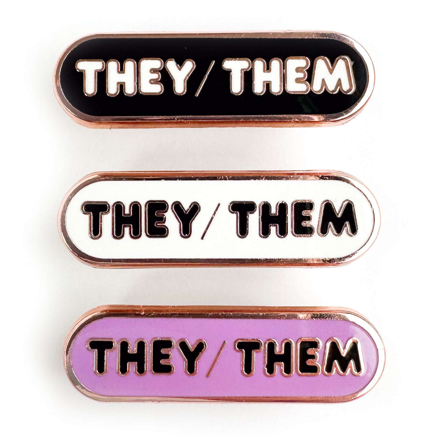Three capsule shaped pins that all have the words "They/Them" on them, one pin is black, one is white, and one is lavender.