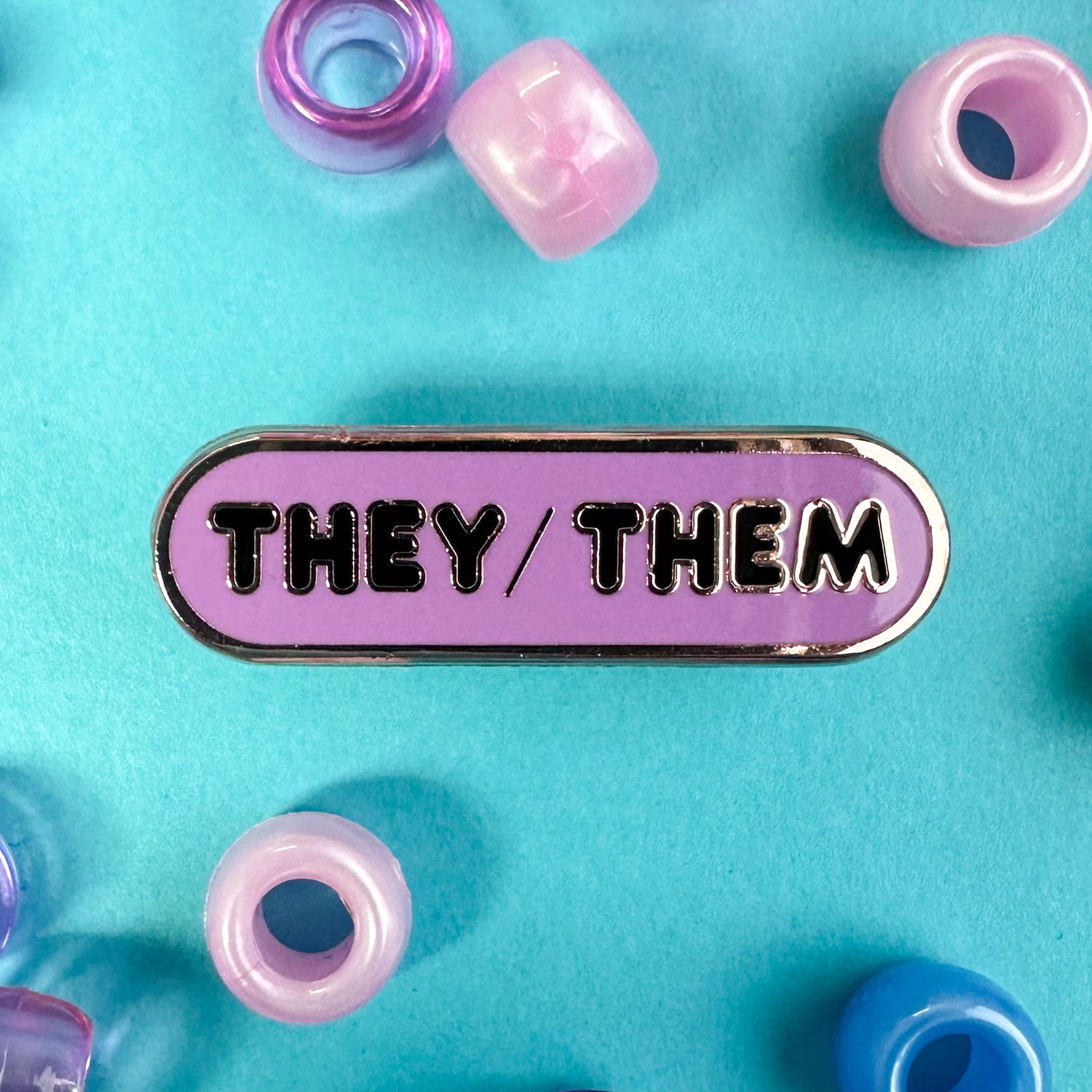 An oval lavender pin with black bubble letters on it. The pin is on a blue background with pink and blue pony beads around it.