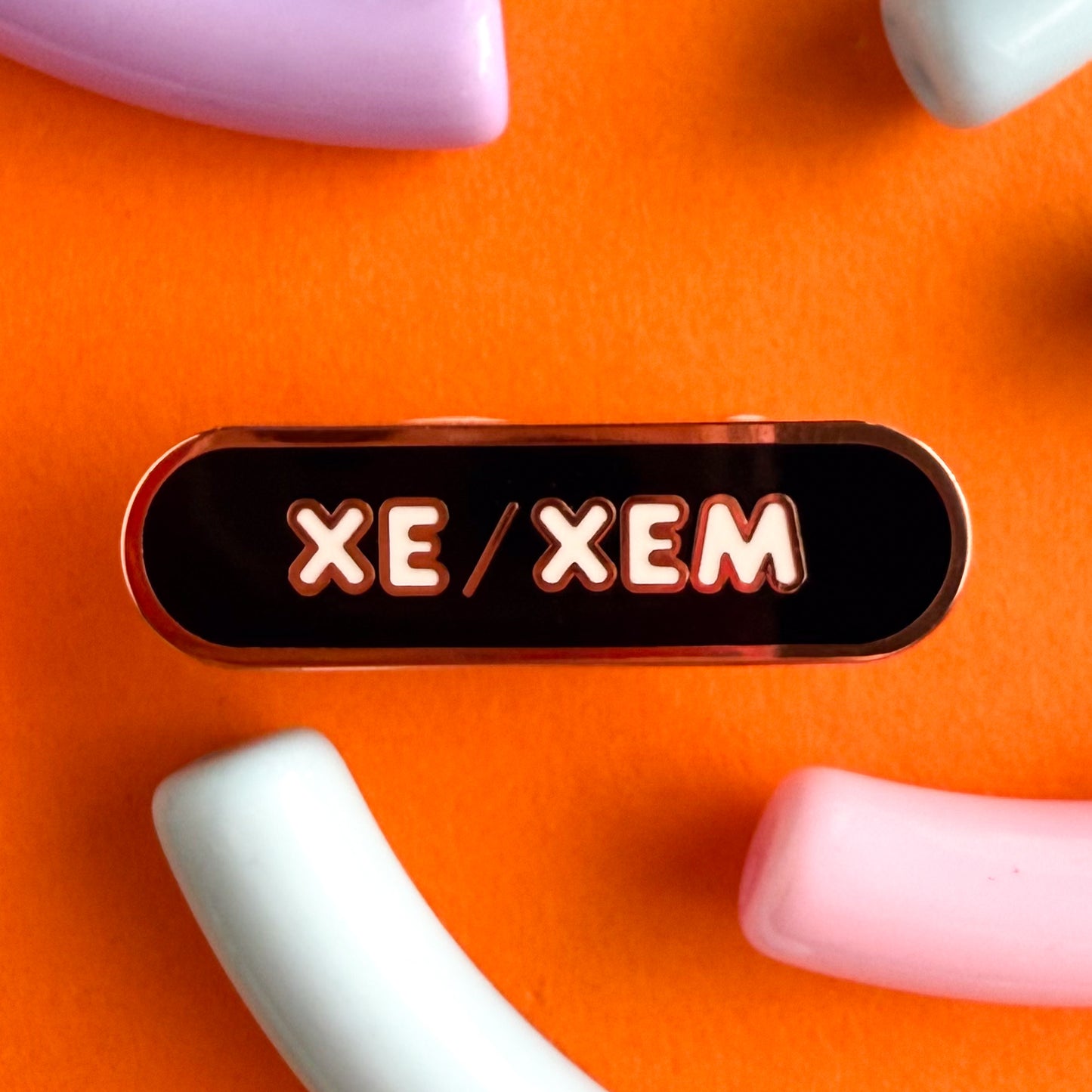 A bandaid shaped pin with the text XE/XEM in bubble letters. The background is black and the text is white. The pin is on an orange background.