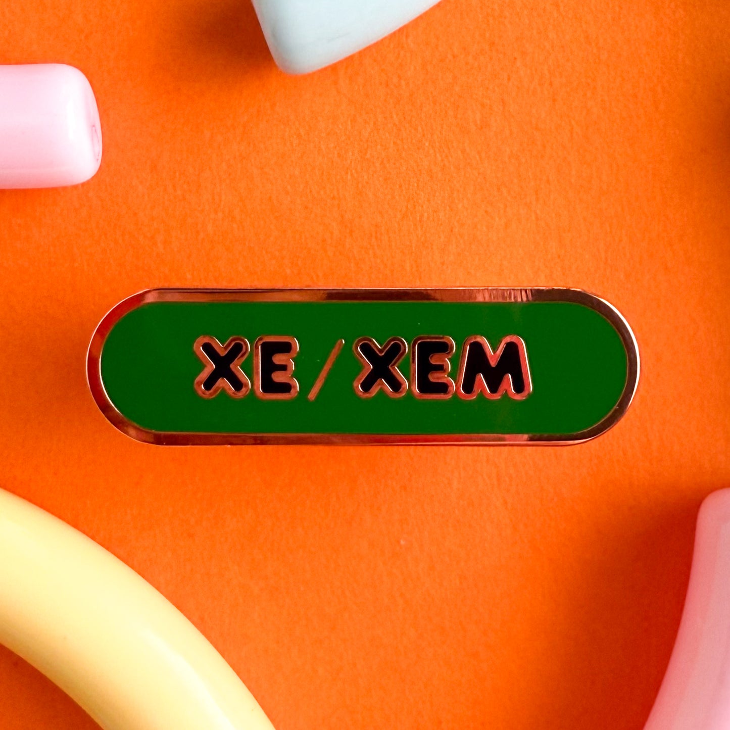 A bandaid shaped pin with the text XE/XEM in bubble letters. The background is green and the text is black. The pin is on an orange background.