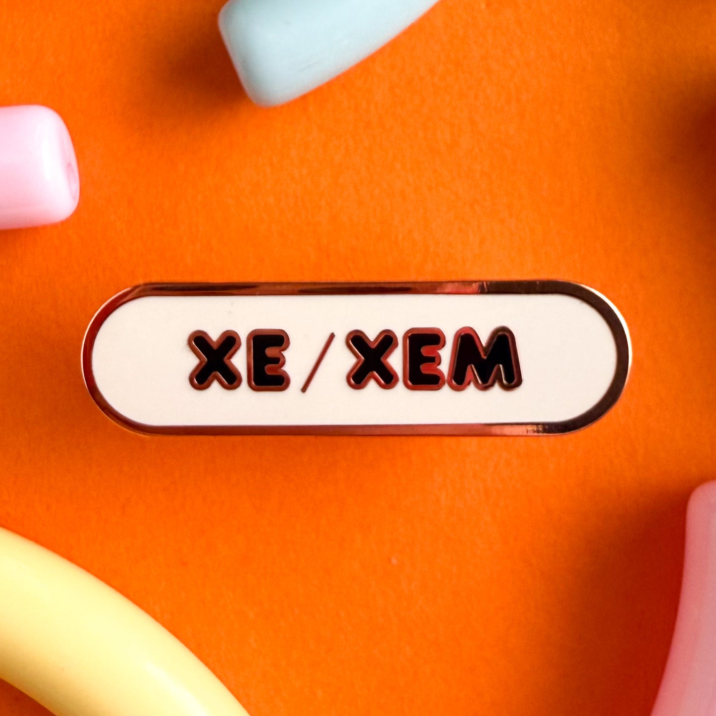 A bandaid shaped pin with the text XE/XEM in bubble letters. The background is white and the text is black. The pin is on an orange background.
