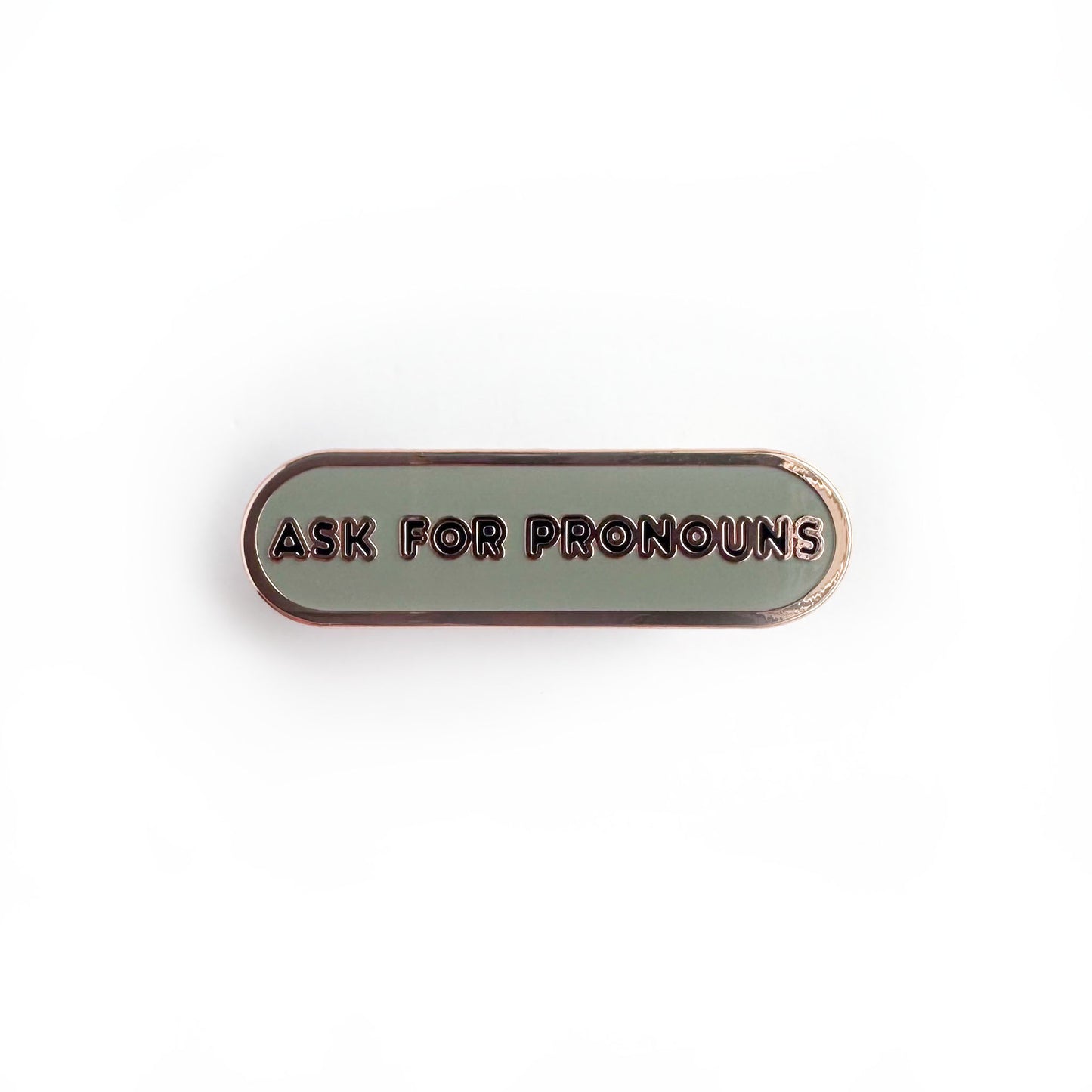 Oval shaped enamel pin with "ask for pronouns" written in black text on a grey background.