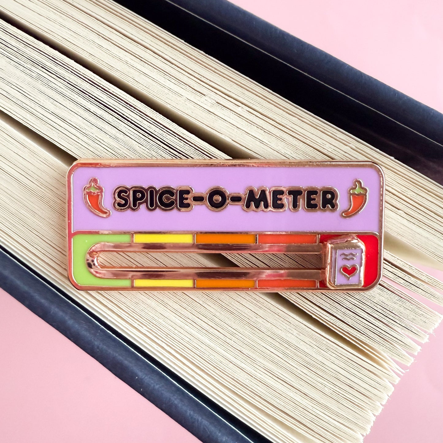 An enamel pin with the title SPICE-O-METER written in black on a pink background.There are two red hot peppers, one before and one after the title. Below the title is a scale with a slider in the middle. The scale is green, yellow, orange, red orange, and red. The slider is a small pink book with a red heart on the cover. The pin is displayed on the splayed edge of a book.