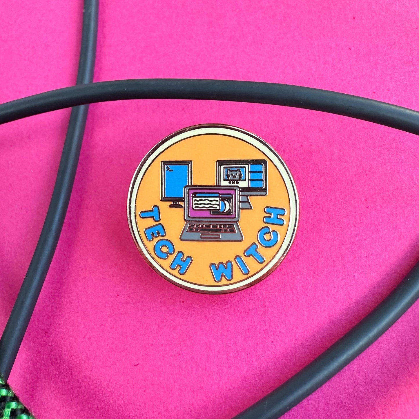 A circular Tech Witch enamel pin on a hot pink background with cords surrounding it. The pin has an image of a laptop surrounded by monitors on it in a fun orange, hot pink, yellow and blue color palette.
