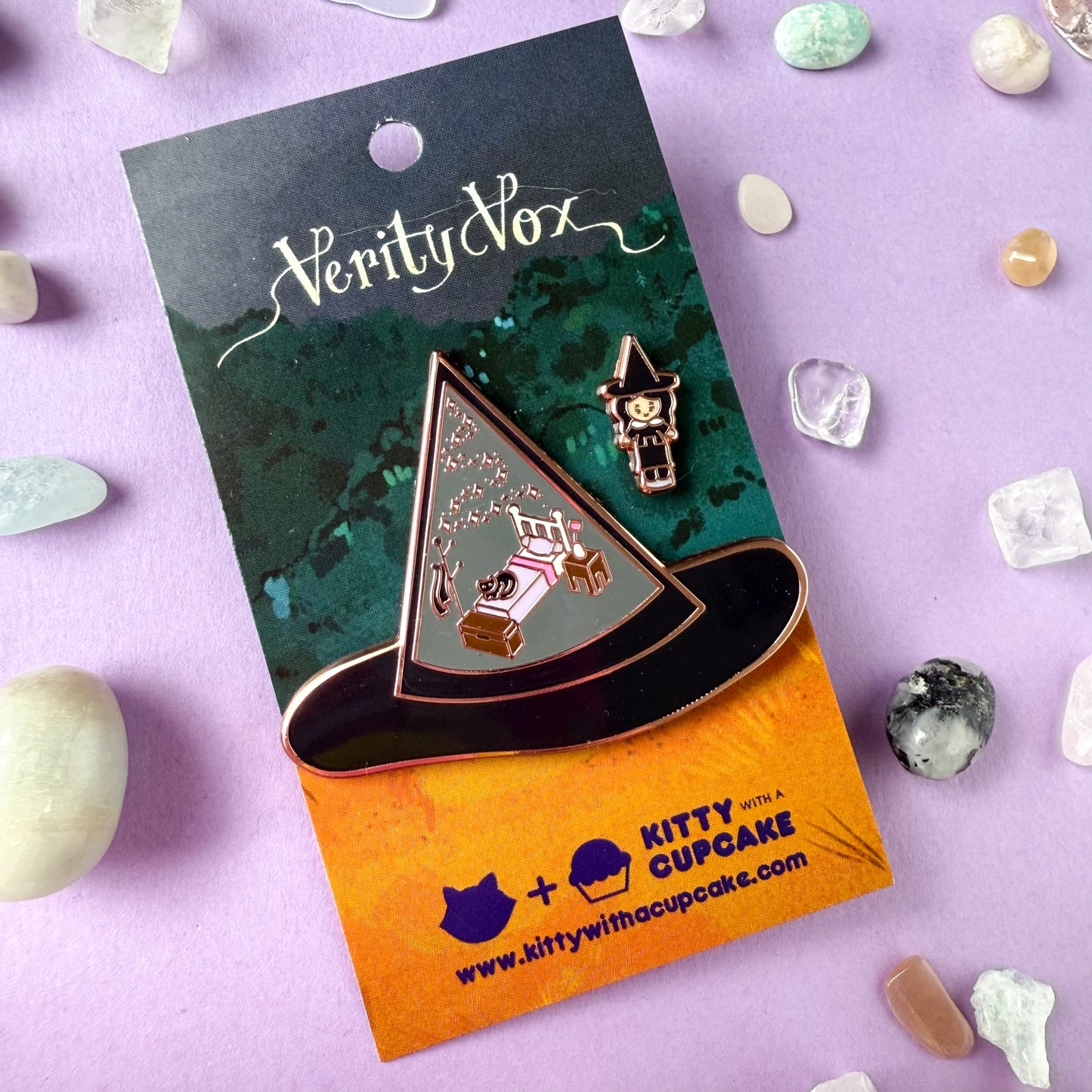 An enamel pin set, one is a large pin shaped like a witch hat with illustrations of a bedroom set and a cat in it. The other pin is a mini pin of a witch wearing a hat that matches the other hat. The pins are packaged on a Verity Vox branded card and are surrounded by small crystals.