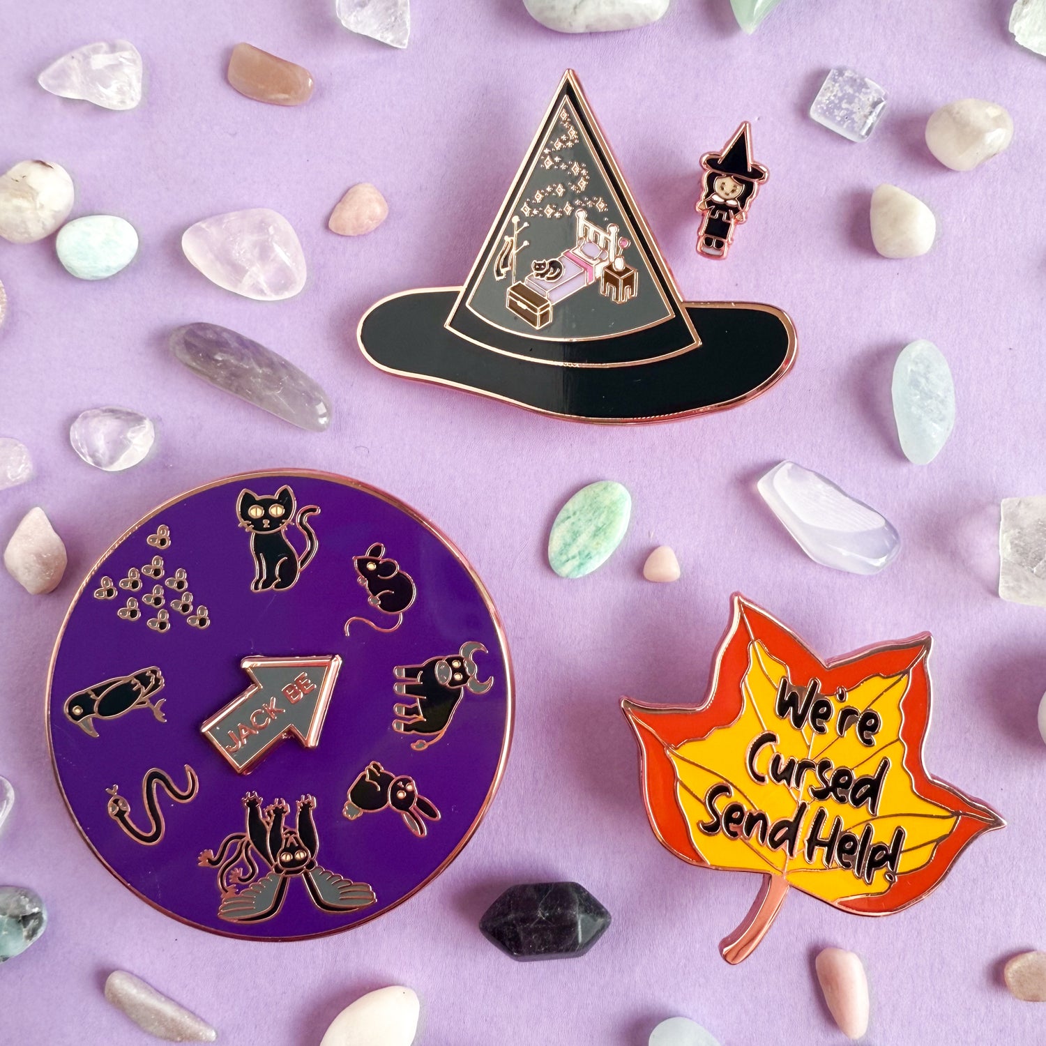 A group of enamel pins on a purple background with crystals around them. There is a circle pin with different animal illustrations on it, there is also a fall leaf, and a witches hat with a tiny witch.