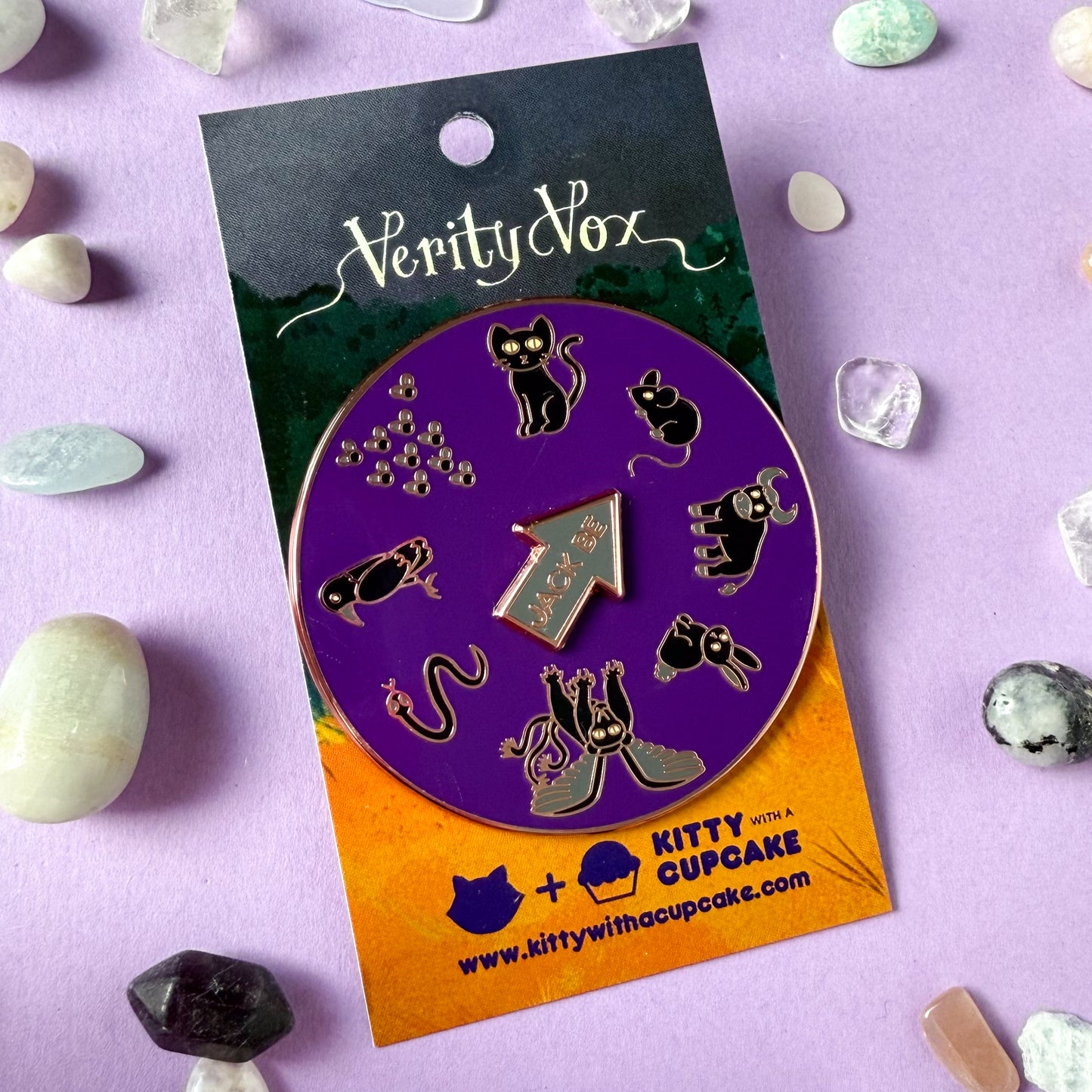 A round purple enamel pin with illustrations of black animals in a circle. There is a black cat, mouse, bull, rabbit, chimera, snake, raven, and swarm of flies. There is a spinning arrow in the middle of the circle that reads "Jack Be". The pin is packaged on a Verity Vox branded card. There are crystals strewn around the card.