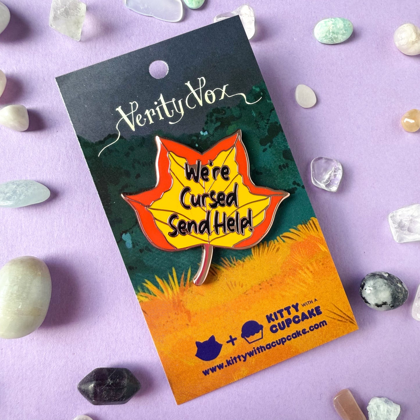 A pin shaped like a tulip poplar leaf in fall colors with the words "We're cursed send help!" written on it. The pin is packaged on a card that reads "Verity Vox" with the Kitty With A Cupcake logo below. There are small crystals strewn about the card.