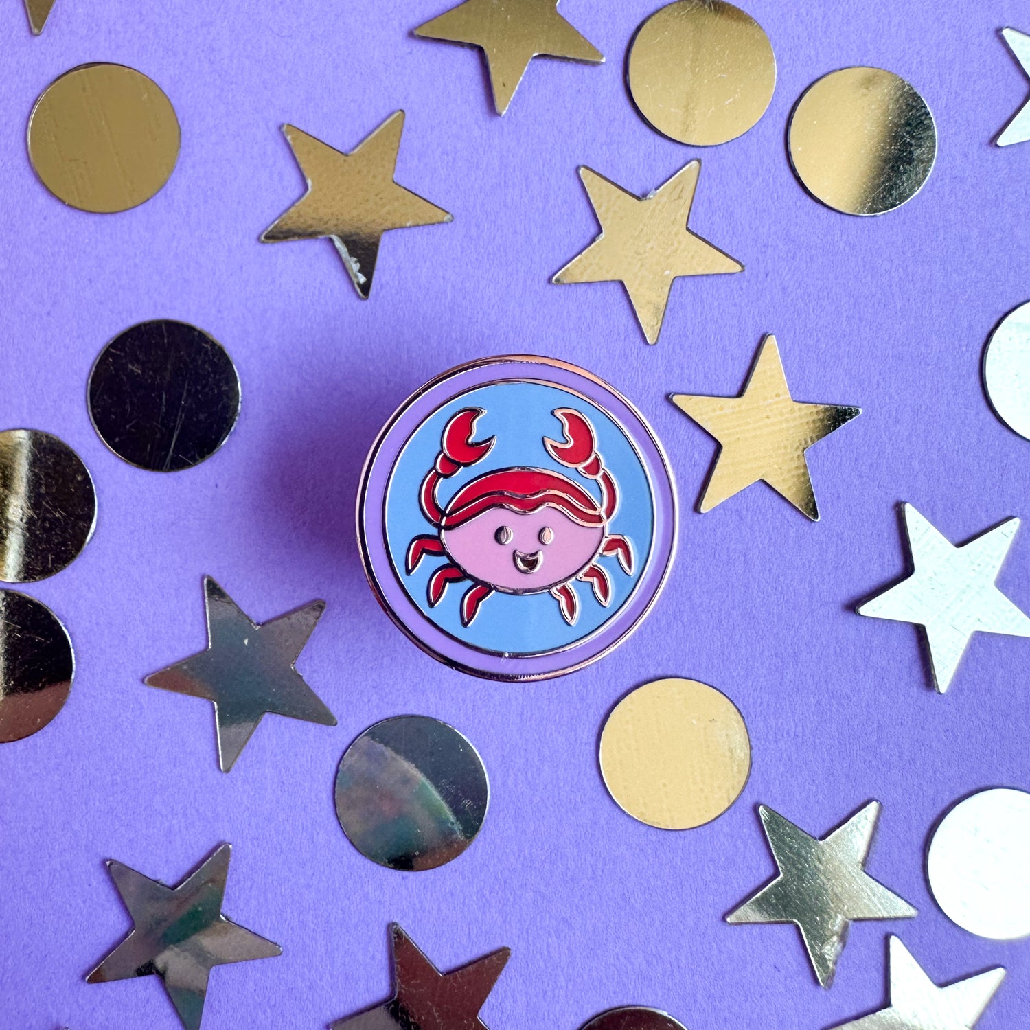 An enamel pin in the shape of a circle with a cute little crab illustration inside of it to represent Cancer in the zodiac. The pin is on a purple background with gold stars and circles around it.