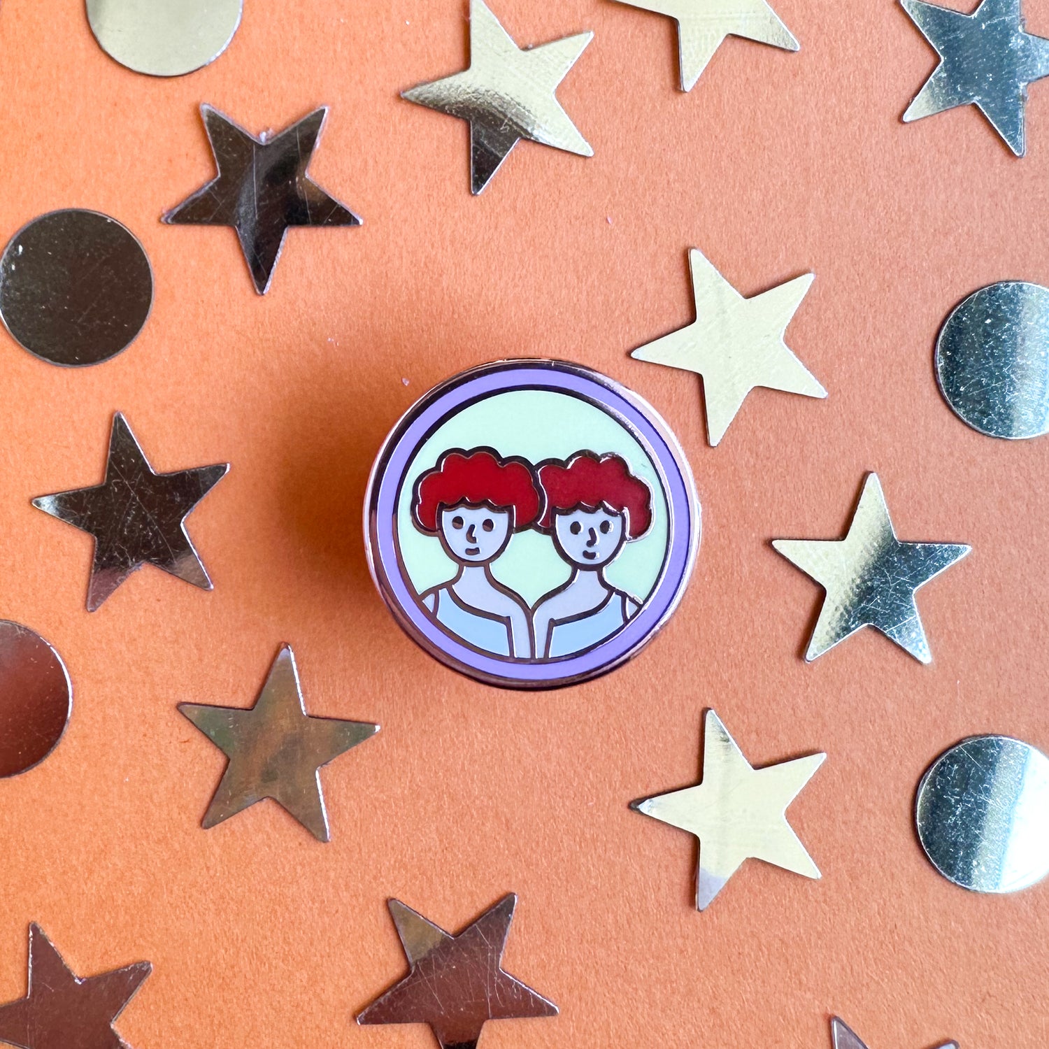 A circular enamel pin with the symbol for Gemini on it, two twins with red hair. The pin is on an orange background with gold stars and circles around it.