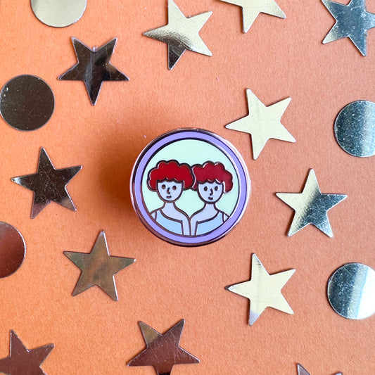 A circular enamel pin with the symbol for Gemini on it, two twins with red hair. The pin is on an orange background with gold stars and circles around it. 