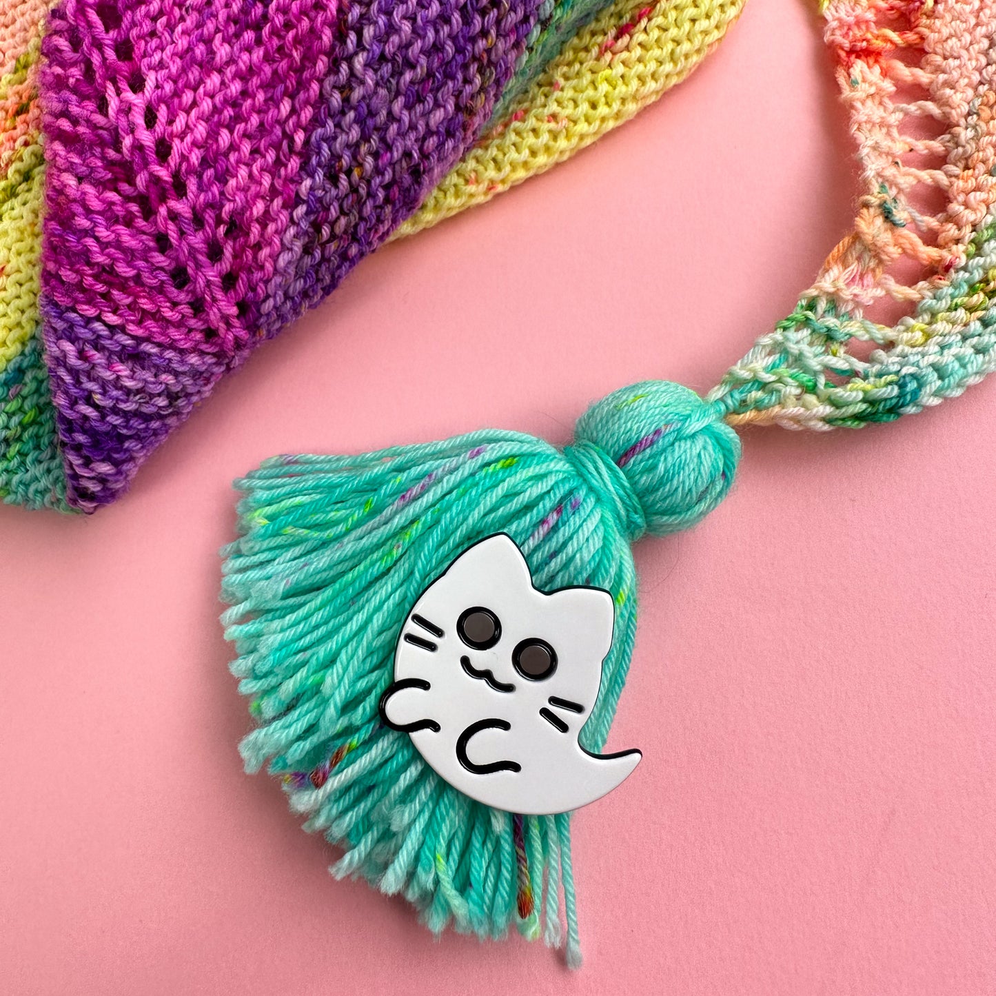 A ghost kitty hair clip attached to a turquoise handmade tassel that is attached to a rainbow hand knit shawl in the corner of the image.