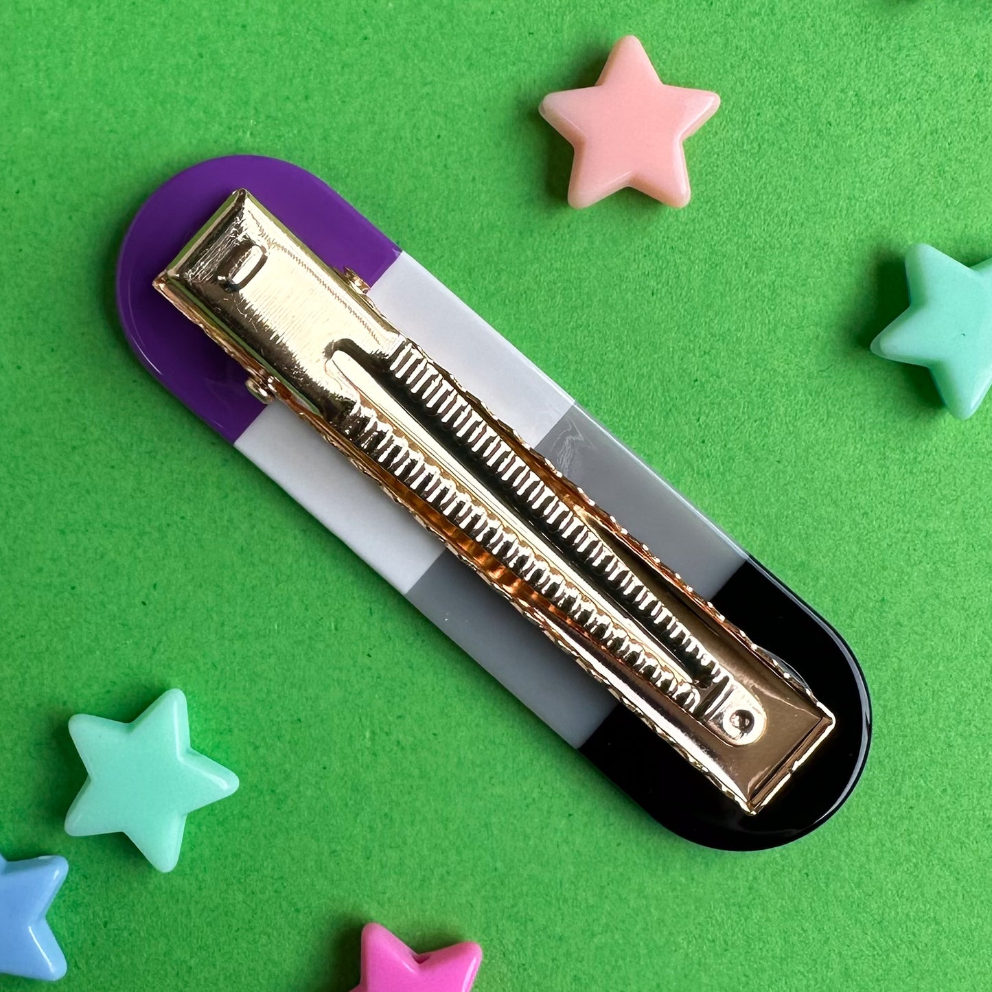 An alligator hair clip with the gold clip part facing up. The clip is oval shaped in the stripes of the Asexual pride flag. It is sitting on a green piece of paper with pastel star beads around it.