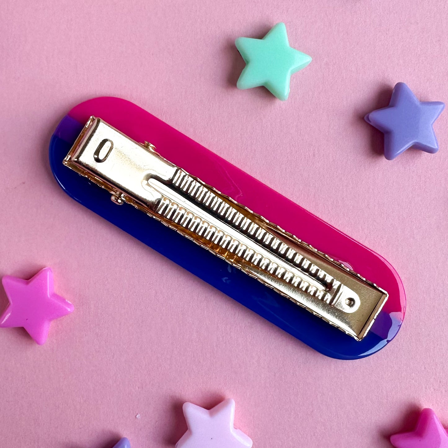 An alligator hair clip flipped up so the gold alligator clip part is on top. The clip is pink, blue and purple. It is sitting on a pastel pink piece of paper with pastel plastic star beads around it.