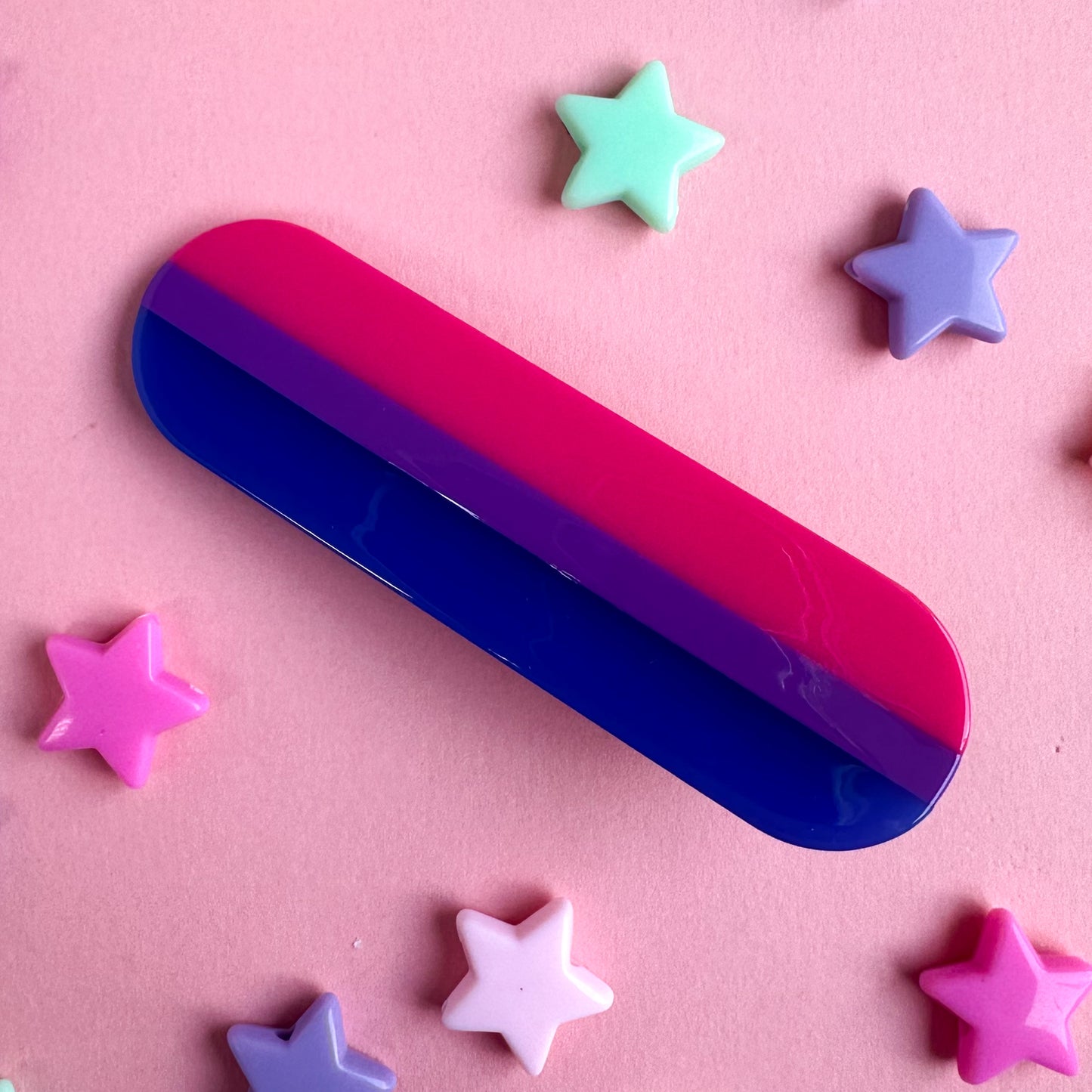 A capsule shaped hairclip in the colors of the bisexual pride flag. The clip is on a pink background with pastel star beads around it.