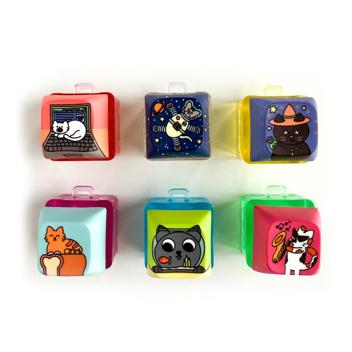 Six square shaped keycaps on clear colorful bases. Each of the keycaps has a cute illustration of a cat on it including a white yelling Laptop Cat, an Astronaut Cat in space, a Witch Cat with a frog familiar, an orange cat on a loaf of bread, a grey cat looking at a goldfish in a bowl, and a calico cat wearing sunglasses and playing the saxophone.