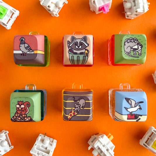 Keycap fidget clickers will illustrations of six different critters on them including a pigeon on the street, a raccoon in a trash can, an opossum with babies on it in a grass field, a squirrel on a pile of acorns on a stump, a rat dragging a slice of pizza up the stairs and a seagull looking angry on top of a container of fries.