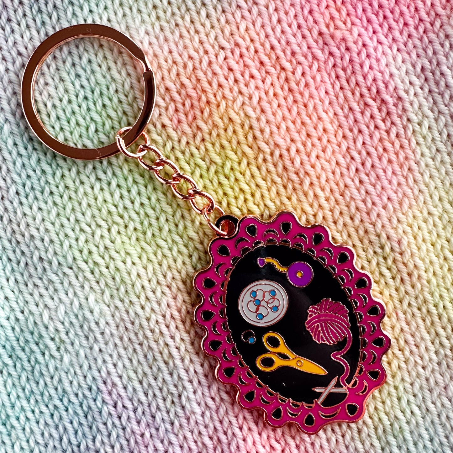 A copper color hard enamel metal keychain with a charm that depicts the perfect knitting project set up with a project on the needles, pink yarn, scissors, stitch markers, and a tape measure.
