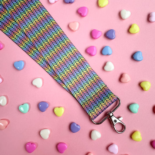 A woven lanyard with a pastel rainbow stockinette stitch graphic printed on it with a silver metal lobster claw clasp at the bottom.  The lanyard is on a pastel pink background with pastel colored heart beads scattered around it. 