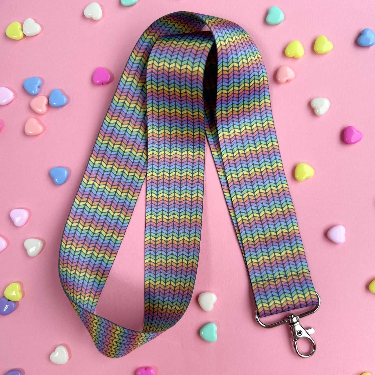 A woven lanyard with a pastel rainbow stockinette stitch graphic printed on it with a silver metal lobster claw clasp at the bottom. The lanyard is bend in half to show how long it is and is on a pink background with pastel heart beads scattered around it. 