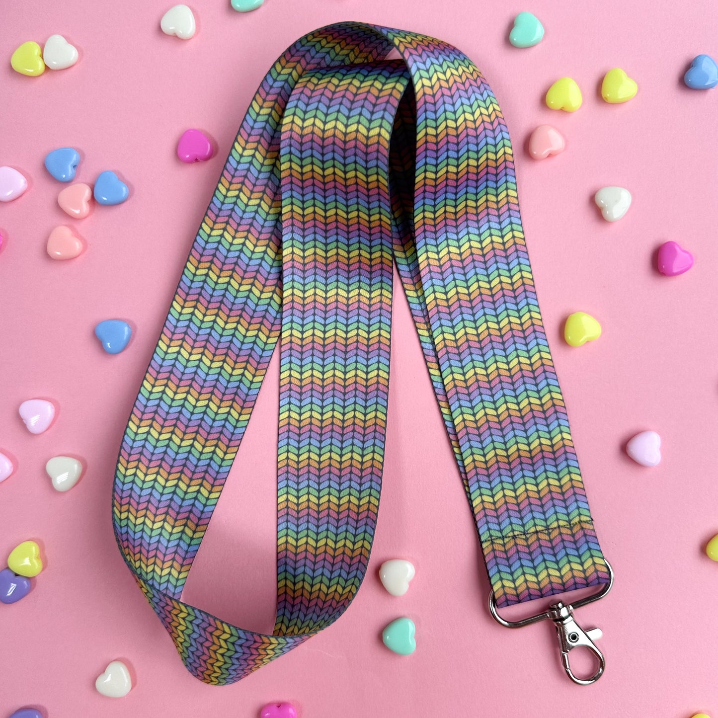 A woven lanyard with a pastel rainbow stockinette stitch graphic printed on it with a silver metal lobster claw clasp at the bottom. The lanyard is bend in half to show how long it is and is on a pink background with pastel heart beads scattered around it.