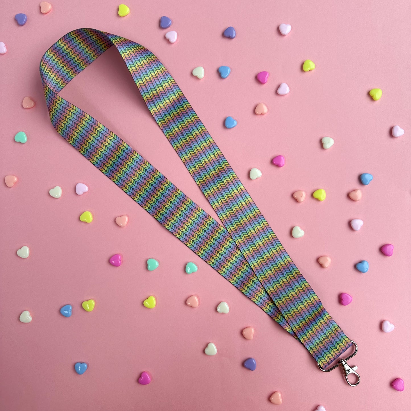 A woven lanyard with a pastel rainbow stockinette stitch graphic printed on it with a silver metal lobster claw clasp at the bottom. This is a further out image showing the entire length of the lanyard on a pastel pink background with pastel colored hearts scattered around it. 