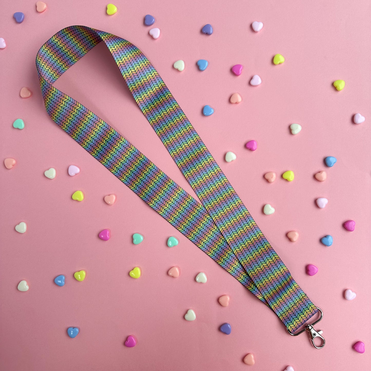 A woven lanyard with a pastel rainbow stockinette stitch graphic printed on it with a silver metal lobster claw clasp at the bottom. This is a further out image showing the entire length of the lanyard on a pastel pink background with pastel colored hearts scattered around it.